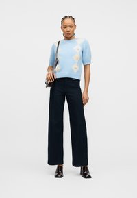 Γυναίκα με πλεξούδες που φοράει ανοιχτό μπλε μπλούζα με μοτίβο και κοντά μανίκια, μαύρο παντελόνι ψηλόμεσο με φαρδιά πόδια, σκουρόχρωμα παπούτσια με τακούνι και τσάντα χιαστί.