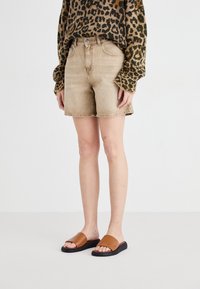Pantalones cortos de mezclilla beige con dobladillo deshilachado, combinados con un suéter marrón de dibujos. Sandalias deslizantes marrones con suela negra. Diseño minimalista.