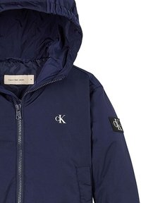 Giacca a vento blu navy in tessuto liscio, con zip frontale, tasche laterali e un logo bianco sul petto con una toppa sulla manica.