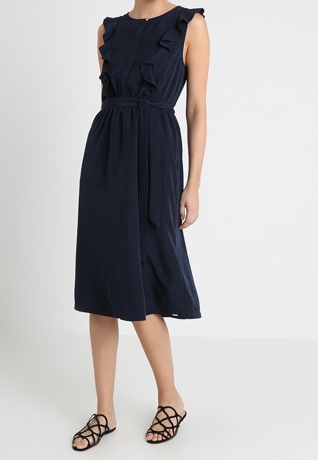 Mörkblå, knälång klänning med volangärmar, ett figurnära midja och ett bälte. Slät tyg med lätt glans, bärs med svarta sandaler.