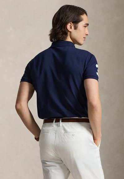 Tengerészkék galléros póló, rövid ujjakkal és fehér részlettel az ujjon; fehér nadrággal és barna övvel párosítva.