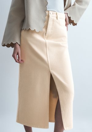 Μπεζ μίντι φούστα με εμπρόσθια σχίση συνδυασμένη με μπεζ σακάκι με κυματιστό τελείωμα και λευκό τοπ, που δείχνει το χέρι και τη μέση του μοντέλου.