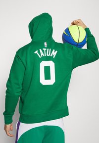 Felpa verde con scritta bianca "TATUM" e numero "0" sulla schiena; persona che tiene un pallone da basket blu e giallo, indossando pantaloni corti multicolori.
