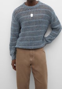 Person in einem blauen offenen Strickpullover und beigen Hosen, mit einer silbernen Anhänger-Halskette, steht mit einer Hand auf der Hüfte.