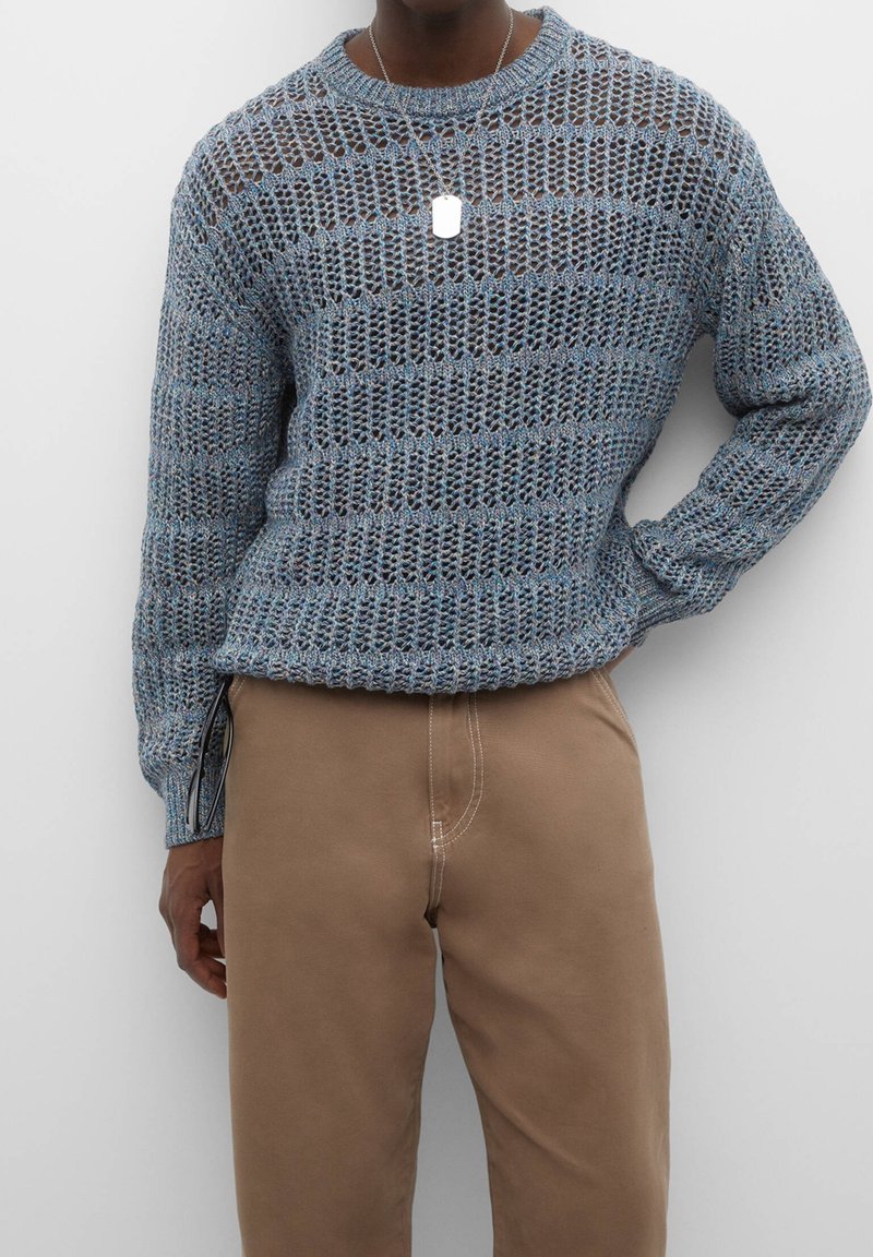 Person in einem blauen offenen Strickpullover und beigen Hosen, mit einer silbernen Anhänger-Halskette, steht mit einer Hand auf der Hüfte.