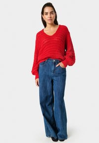 Pulover roșu tricotat cu o gât V larg și mâneci lungi, asortat cu blugi albastri din denim, cu talie înaltă și croi lejer. Se văd pantofi negri, fără toc.
