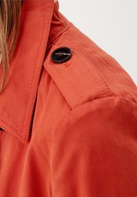 Bavlněný trenčkot ve jasně oranžové barvě s výrazným límcem. Obsahuje černý knoflík na rameni a podrobné švy.