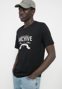 Crna pamučna majica s bijelim i zlatnim natpisom, kratkih rukava i opuštenog kroja. Kombinirana s svijetloplavim trapericama.