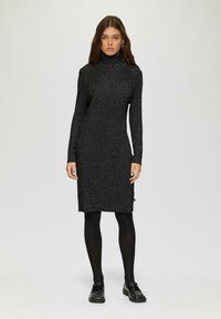 Schwarzes, langärmeliges Kleid mit hohem Kragen, hergestellt aus strukturiertem Stoff mit subtiler Shimmer-Oberfläche. Dargestellt mit schwarzen Strumpfhosen und verzierten Schuhen getragen.