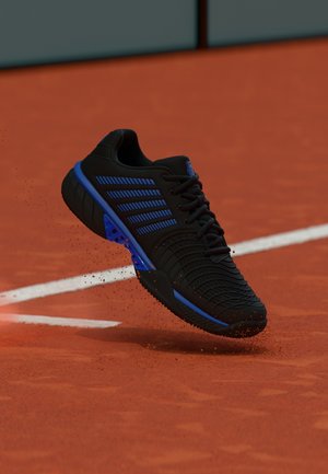 Black and blue tennis shoe kicking up red clay dust on a tennis court near white boundary lines.