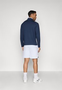 Námornícke polo tričko s dlhým rukávom a bielym golierom, spárované s bielymi šortkami. Medzi vlastnosti patrí rovný strih a hladká textúra látky.