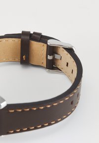Bracelet de montre en cuir brun avec une doublure beige, des coutures blanches et une boucle en métal argenté. Présente des trous de réglage et une texture lisse.