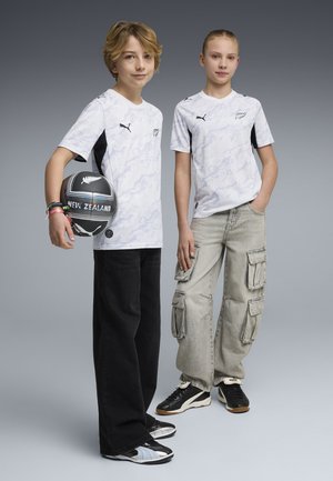 Twee jongeren dragen witte rugbyshirts van Nieuw-Zeeland; een van hen houdt een zwarte rugbybal vast met het opschrift "New Zealand".