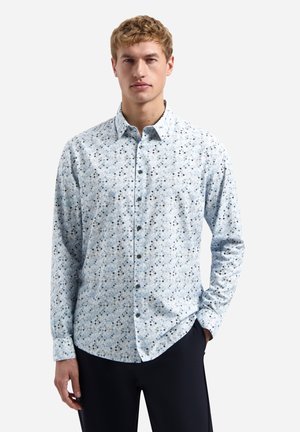 Jonge man die een lichtblauw overhemd met lange mouwen en een kleine donkere geometrische print draagt, zwarte broek, staand met één hand in de zak.