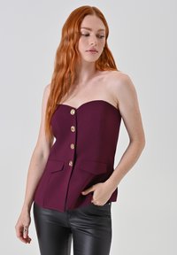 Top senza spalline color bordeaux con una vestibilità sartoriale, caratterizzato da dettagli con bottoni dorati e due tasche anteriori, abbinato a pantaloni di pelle nera.