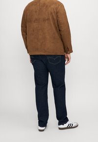 Homme portant une veste en daim marron, un jean bleu foncé et des baskets blanches avec des rayures noires, se tenant contre un fond clair uniforme.