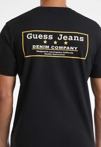 Czarny t-shirt z tyłu z prostokątnym logo w żółtej ramce, na którym widnieje napis "Guess Jeans Denim Company" oraz mniejszy tekst "Zaprojektowane w Los Angeles, Kalifornia."