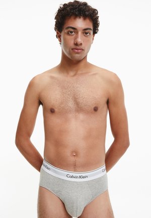 Boxer grigi in cotone con una vita elastica bianca, con il logo Calvin Klein. Modello in piedi con le braccia lungo i fianchi, torso nudo visibile.