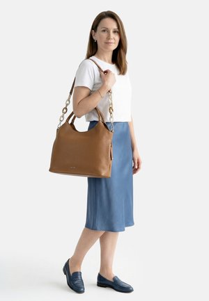 Žena v bílém tričku a modré sukni drží velkou hnědou koženou kabelku s řetízkovým popruhem přes rameno, stojí na bílém pozadí.