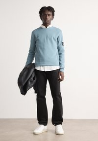 Lys blå genser med rund hals, svarte jeans, hvite sneakers og en svart skinnjakke drapert over armen. Glatt tekstur, avslappet passform.