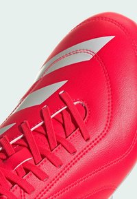 Scarpa da calcio sintetica rossa con finitura testurizzata, caratterizzata da strisce bianche e lacci piatti. Punta arrotondata e dettagli di cucitura rinforzati.