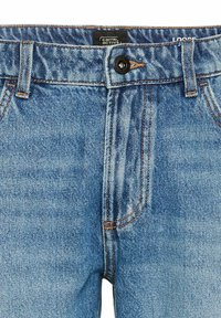 Løse blå denimjeans med en tekstureret finish, prydet med orange syninger og en sort knaplukning. Mærket viser "Camel Active."