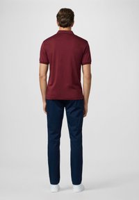 Burgundská polo košile s krátkým límečkem a tmavě modrými kalhotami. Košile má kontrastní lemování a model má na sobě bílé boty.