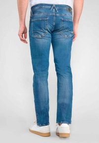 Jean en denim bleu avec une coupe droite, avec des poches arrière et une étiquette de taille visible. Portés avec des baskets blanches.