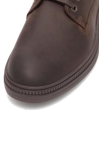 Hnedá kožená obuv s zaoblenou špičkou, jemnou textúrou a ryhovanou gumovou podrážkou s dizajnom na šnurovanie.