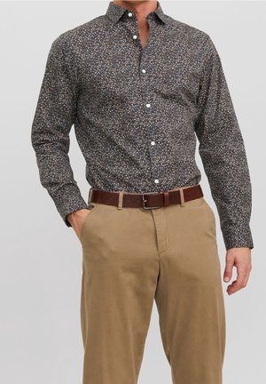 Mann trägt dunkles, langärmeliges Hemd mit floralem Muster, khakifarbene Hose und braunen Ledergürtel, eine Hand in der Tasche vor schlichtem Hintergrund.