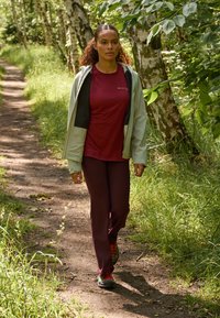 Bordeaux sportshirt, lichtgroene jas en bordeaux broek. Zwarte en rode schoenen. Omringd door groen op een onverharde weg.