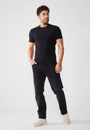 Svart kortermet t-skjorte, slim fit, kombinert med mørke jeans og lyse sneakers, modellert av en mann med krøllete hår mot en nøytral bakgrunn.