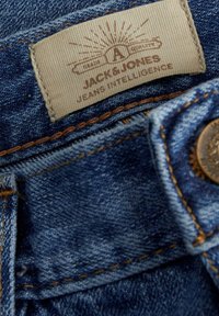 Jeans in denim blu con un'etichetta in tessuto che presenta "JACK&JONES" e "JEANS INTELLIGENCE", accentuati da cuciture arancioni a contrasto e un bottone in metallo.