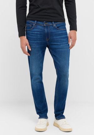 Jeans de denim azul ajustados con un lavado sutil, cinco bolsillos y costuras tradicionales. Combinados con zapatillas blancas y una camisa negra de manga larga.