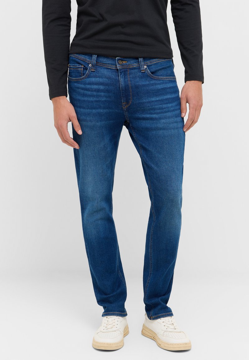 Keskeny szabású kék denim farmer, finom mosással, öt zsebbel és hagyományos varrással. Fehér sportcipővel és fekete hosszúujjú inggel párosítva.