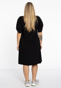Schwarzes knielanges Kleid mit Puffärmeln, glatter Textur und entspanntem Schnitt. Kombiniert mit silbernen Sportschuhen.