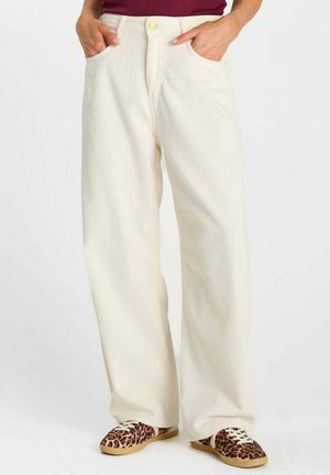 Persona che indossa pantaloni di velluto a coste color avorio a gamba larga, top bordeaux e sneaker con stampa leopardata e lacci bianchi, mani nelle tasche.