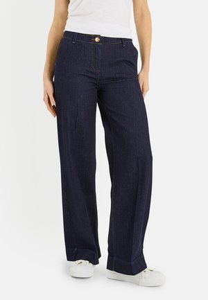 Mujer con vaqueros de pierna ancha azul oscuro con botones delanteros, combinados con zapatillas blancas y una camiseta blanca, posando frente a un fondo liso.