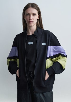 Vrouw draagt een zwarte jas met rits en paarse en groene strepen op de mouwen, staat tegen een effen grijze achtergrond met haar handen in de zakken.