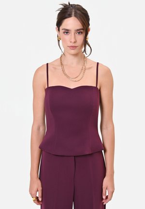 Donna con capelli castani raccolti in uno chignon basso, indossa un top viola scuro senza maniche e pantaloni coordinati, accessoriata con orecchini a cerchio dorati e collane a più strati.