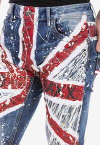 Jean en denim déchiré avec un design Union Jack, dans des couleurs rouge, blanc et bleu, détails de peinture éclaboussée, et un bouton en forme de crâne.
