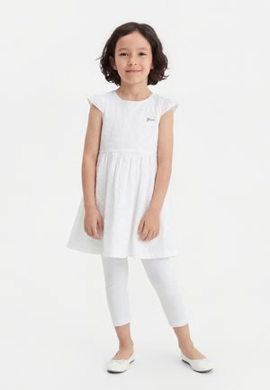 Fată tânără cu păr scurt și închis la culoare, purtând o rochie albă fără mâneci, colanți albi și balerini albi, în picioare pe un fundal simplu.