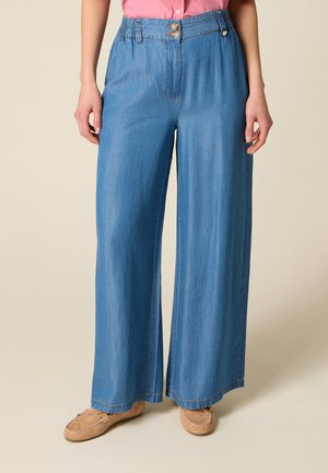 Kobieta nosząca jasnoniebieskie, szerokie dżinsy z podwójnymi drewnianymi guzikami i jasnobrązowe mokasyny, stojąca na beżowym tle.