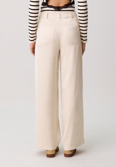 Vue de dos d'une personne portant un pantalon larges crème, un haut à manches longues rayé, des chaussures en daim beige et un soutien-gorge en dentelle noire.