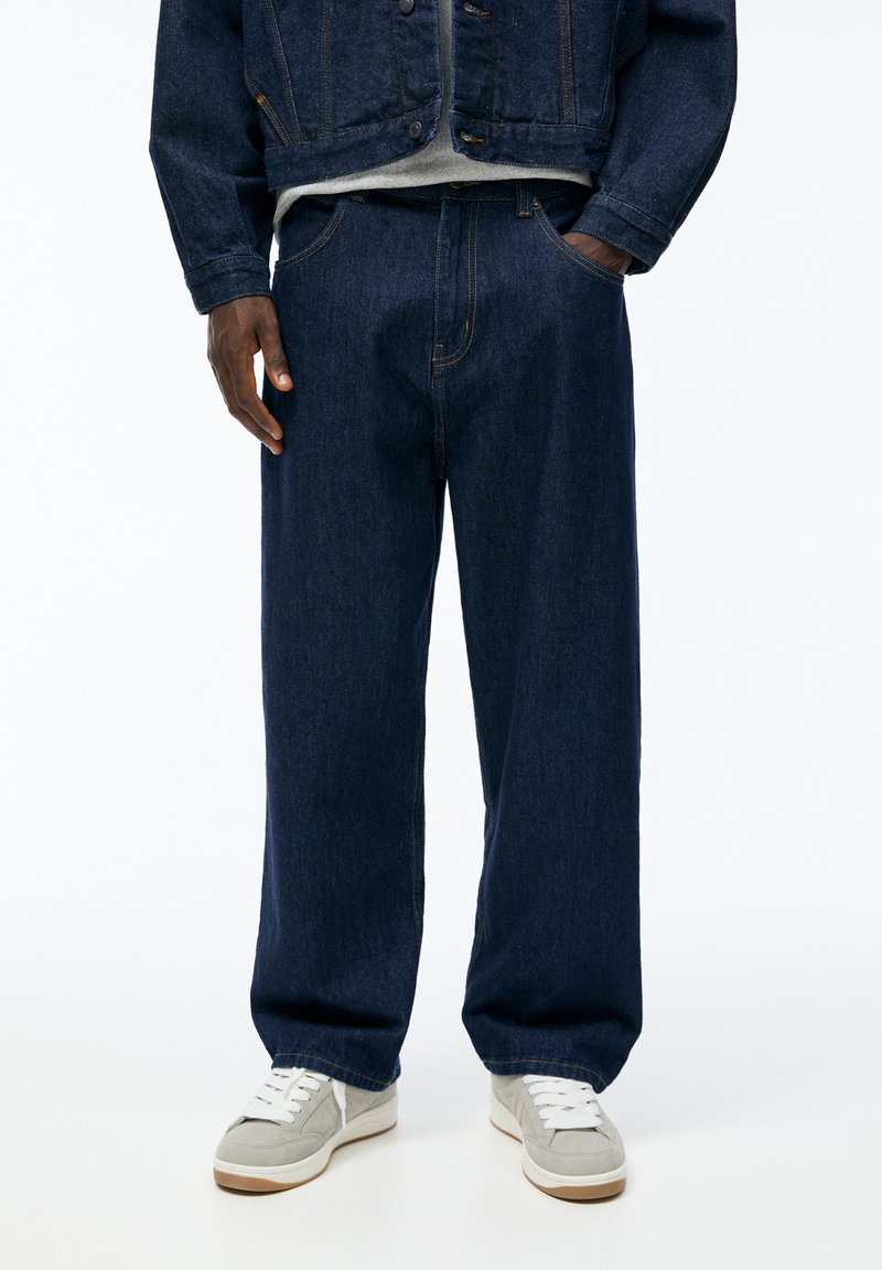 Mørkeblå denim jeans med en afslappet pasform, der har kontrasterende syninger og et klassisk fem-lomme design, parret med grå sneakers.