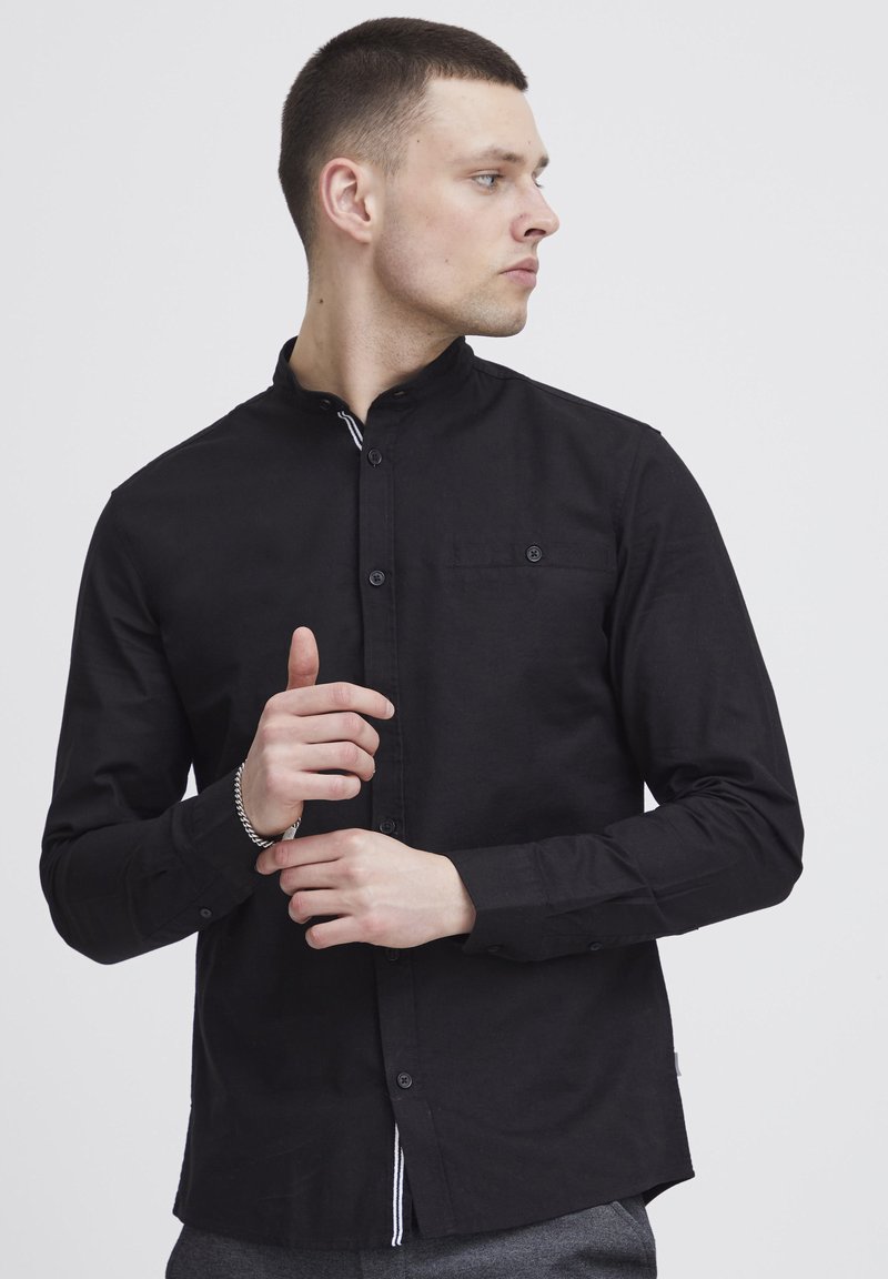 Camicia nera con colletto a band, maniche lunghe, tasca frontale e un accento a righe discreto all'orlo. Tessuto liscio, design aderente.