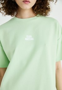 Svetlozelené bavlnené tričko s okrúhlym výstrihom a krátkymi rukávmi, na prednej strane s bielym nápisom "CLUB MATCHA".