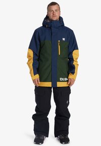 Chaqueta de nieve en azul marino, verde y amarillo con un bolsillo con cremallera y detalle del logotipo. Combinada con pantalones y zapatos negros. Material impermeable.
