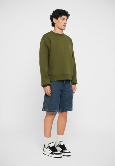Felpa verde oliva con una vestibilità ampia, abbinata a pantaloni shorts di jeans blu. Il modello indossa calzini bianchi fino alla caviglia e sneakers bianco e nere.