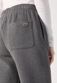 Pilkos sportinės kelnės, pagamintos iš tekstūruoto audinio, su elastiniu juosmeniu ir galiniu kišeniu, kuriame yra žalias „Lacoste“ krokodilo logotipas.